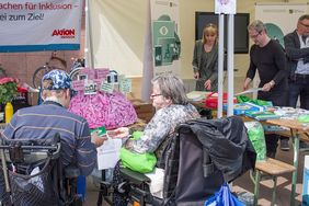 Zwei Rolli-Fahrer sitzen vor einem Gehirn aus Pappmaché an einem Stand. Im Hintergrund ist der ehemalige Behindertenbeauftragte von Sachsen, Stephan Pöhler, zu sehen.