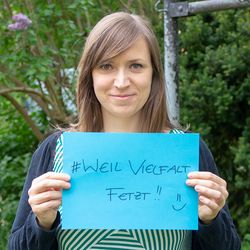 Kerstin Helm steht in einem Garten und hält ein blaues Blatt Papier mit den Worten # Weil Vielfalt fetzt in die Kamera. Sie lächelt.
