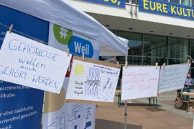 Einige Protestplakate hängen zwischen den Ständen. Auf ihnen steht zum Beispiel: Gehörlose wollen auch gehört werden.